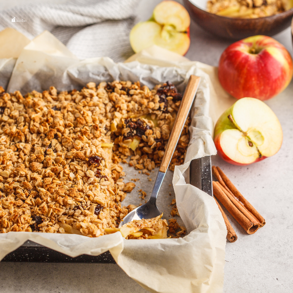 A freshly baked apple crumble in a parchment-lined baking dish, with a spoonful taken out, surrounded by apples and cinnamon sticks.