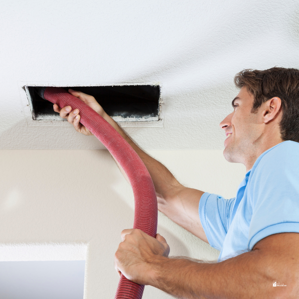 A man in a blue shirt uses a red vacuum hose to clean an air duct in the ceiling of a home.