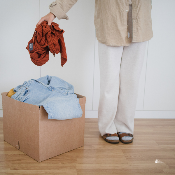 A person holding clothing while standing next to a box filled with folded jeans and other items ready for donation or organization.