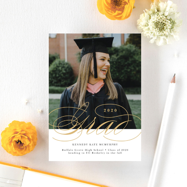 A graduation announcement card featuring a smiling young woman in a cap and gown, placed on a table with flowers and a pencil.