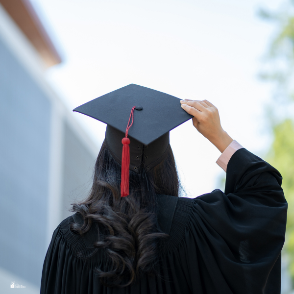 A graduate wearing a black cap and gown, seen from behind while adjusting their mortarboard tassel outdoors.