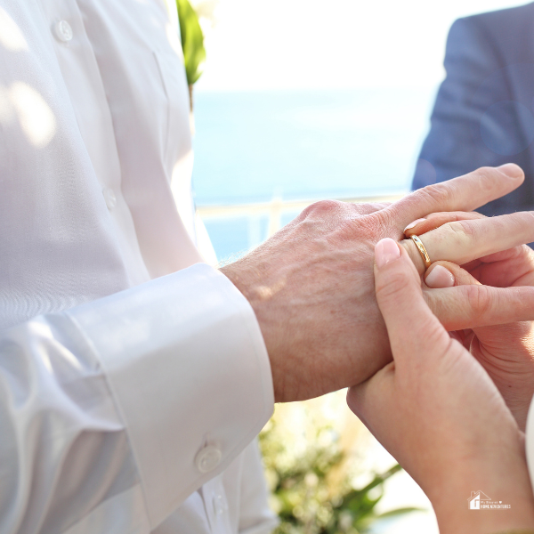 A close-up of a couple exchanging wedding rings during an outdoor ceremony with a scenic ocean backdrop.