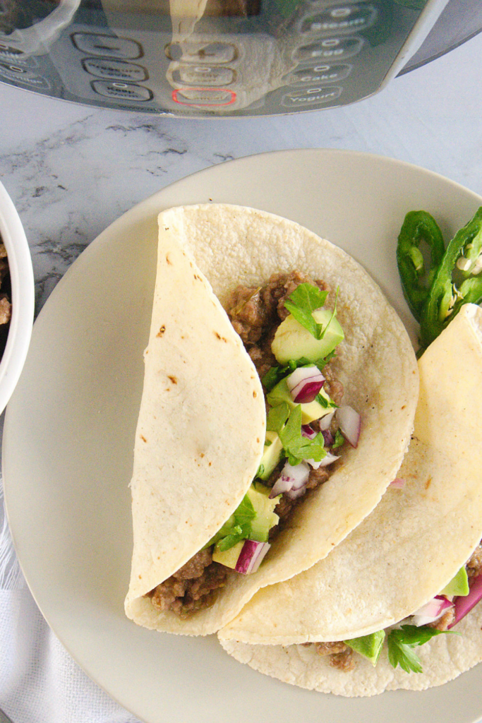 Two soft corn tortillas filled with pressure cooker salsa verde beef, topped with avocado, red onion, and cilantro, served on a plate with sliced jalapeños and an Instant Pot in the background.