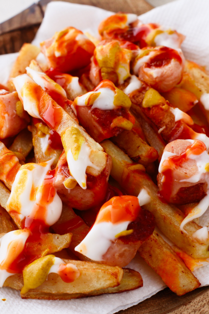 A plate of Salchipapas featuring golden fries topped with sliced hot dogs, ketchup, mayonnaise, mustard, and aji sauce, showcasing a vibrant and flavorful Peruvian street food dish.