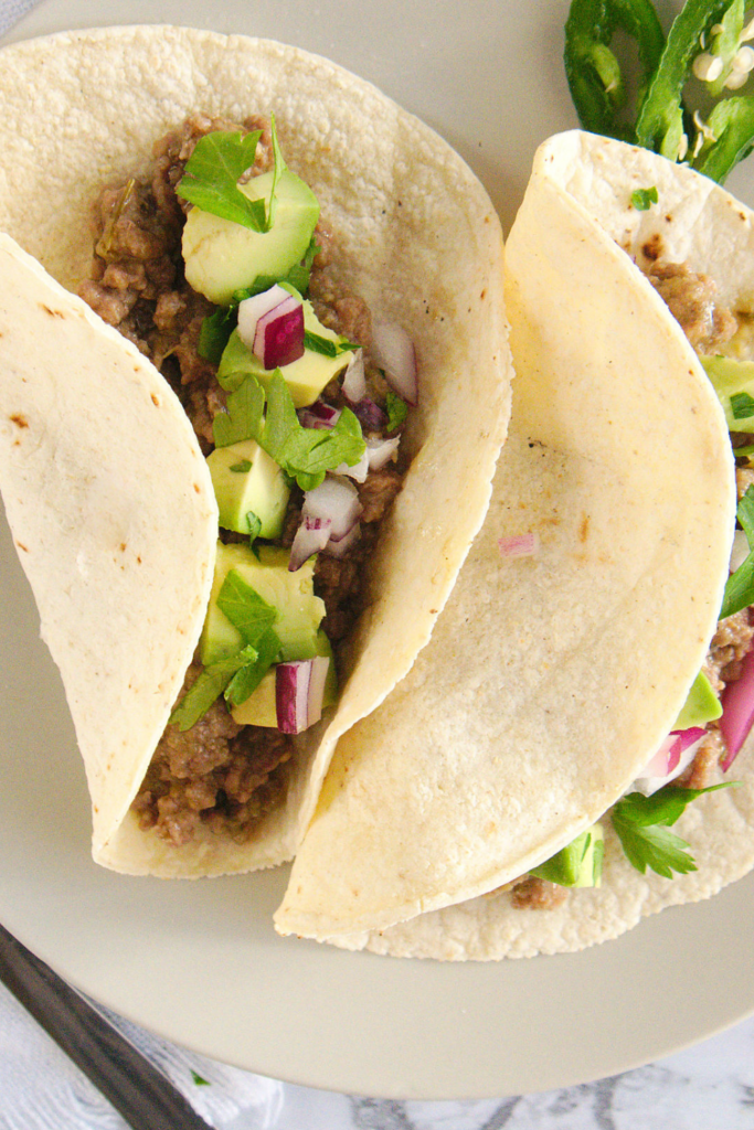 Two corn tortillas filled with pressure cooker salsa verde beef, topped with diced avocado, red onion, and fresh cilantro, served on a plate with sliced jalapeños.