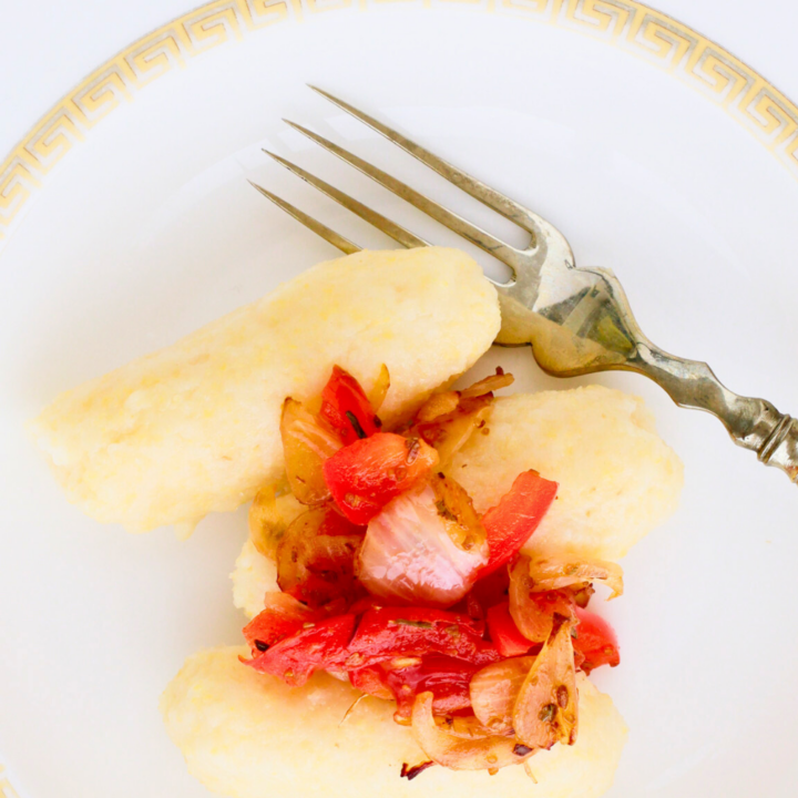 A close-up of Puerto Rican guanimes served with sautéed onions, tomatoes, and peppers on a decorative white plate, highlighting the traditional cornmeal dumplings' soft texture and rich flavors.