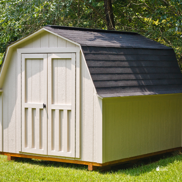 A beige barn-style storage shed with a black shingled roof sits on a well-maintained lawn, surrounded by green trees in the background.