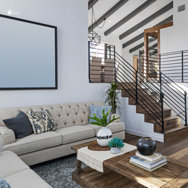 A bright and airy living room with a neutral-toned tufted sofa, rustic wooden coffee table, and modern staircase featuring black metal railings and exposed wood steps.