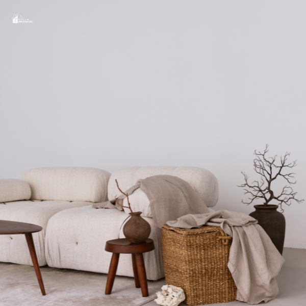 A minimalist living space with a soft, cream-colored couch, textured throw blanket, woven basket, and earthy decor against a simple white wall.