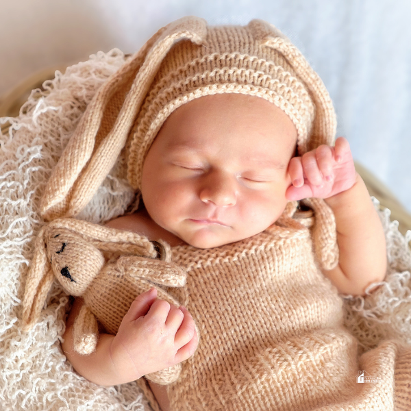 A sleeping newborn baby dressed in a knitted bunny outfit, cuddling a matching bunny toy while resting on a soft blanket.