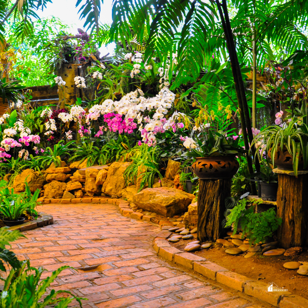 A tropical garden pathway lined with blooming orchids, ferns, and decorative pots, leading through a lush, green oasis with natural stone and brick elements.