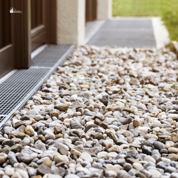 A close-up view of a French drain system with small, smooth gravel stones surrounding a metal drainage grate near the foundation of a house.