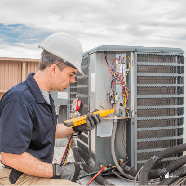 A professional HVAC technician wearing a hard hat and gloves uses a multimeter to check the wiring of an outdoor air conditioning unit on a rooftop.