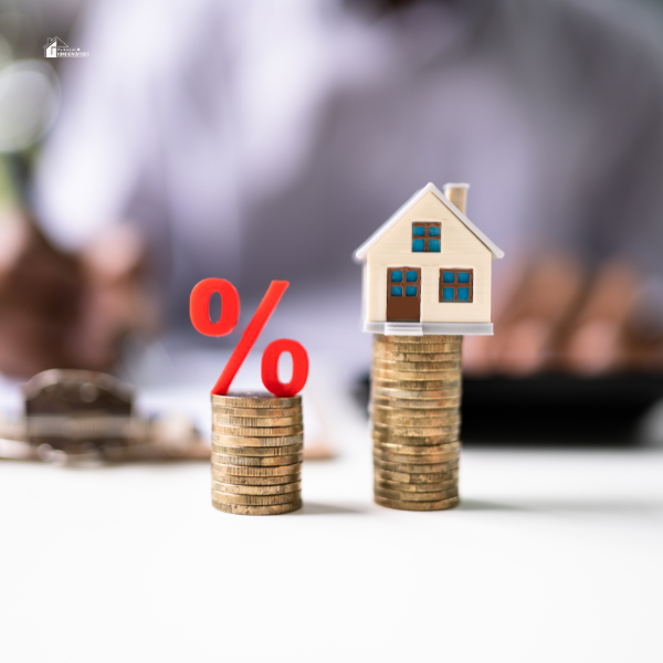 A small model house sits atop a stack of coins next to a red percentage symbol on a smaller coin stack, representing real estate investment and loan interest rates.