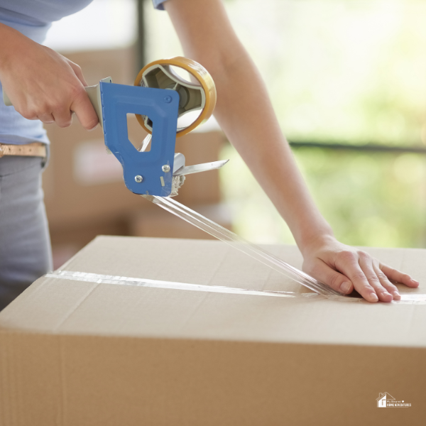 A close-up of a person sealing a moving box with a packing tape dispenser, ensuring the box is securely closed.