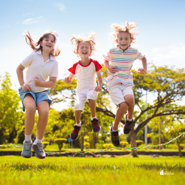 Three children joyfully jump rope together in a lush green park under a bright blue sky.