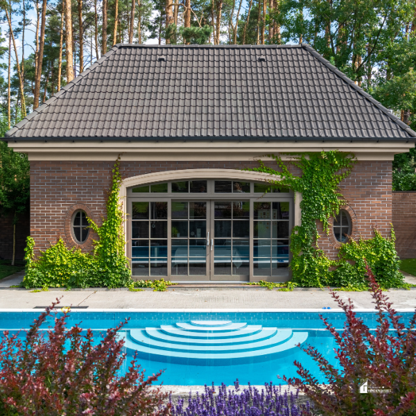 A modern poolside area with a brick pool house surrounded by lush greenery and colorful plants.