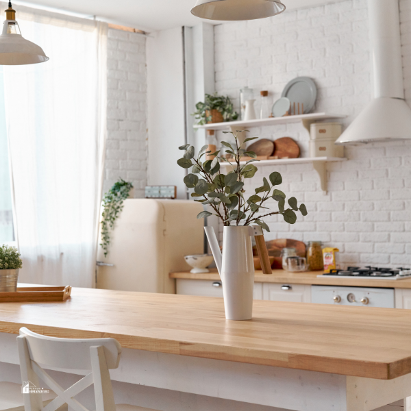 A cozy farmhouse-style kitchen with a wooden dining table, white brick walls, open shelving, and a large window allowing natural light to brighten the space, creating a welcoming and homey environment.