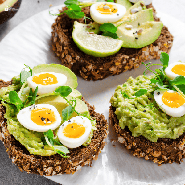 Whole grain toast with avocado spread, boiled eggs, lime slices, and fresh greens on a white plate.