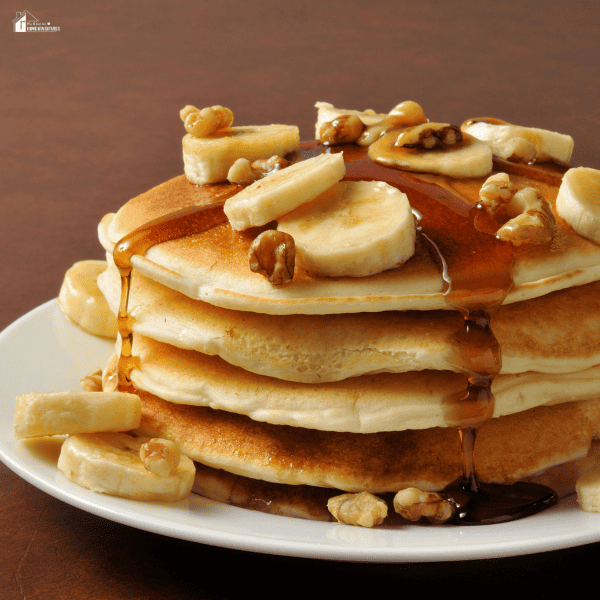 A plate of fluffy pancakes topped with banana slices, walnuts, and drizzled with syrup, served on a white dish.