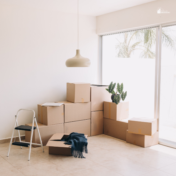 A professional mover wearing a red cap and blue overalls stacking cardboard boxes onto a hand truck in a bright room.