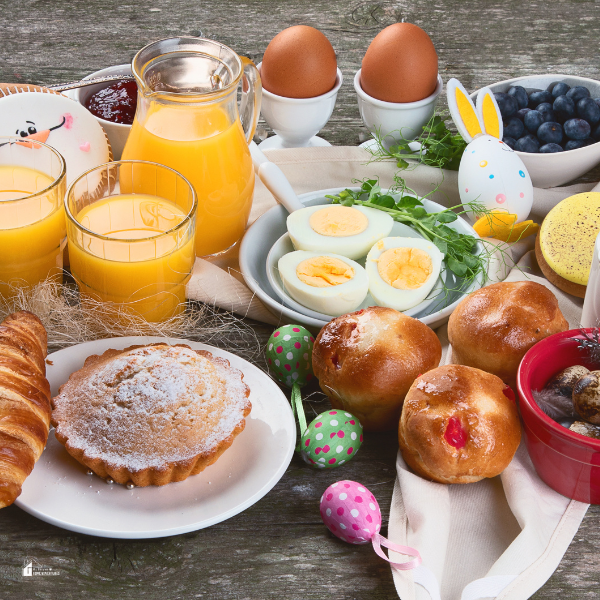 A rustic Easter brunch table filled with soft-boiled eggs, pastries, fresh fruit, and orange juice, decorated with colorful Easter eggs and a bunny figurine.