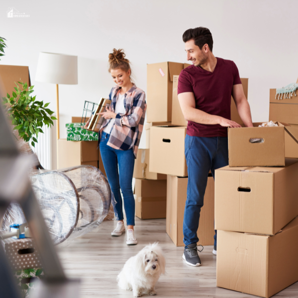 A couple prepares for a move by organizing and packing cardboard boxes in their living room with their small white dog nearby.