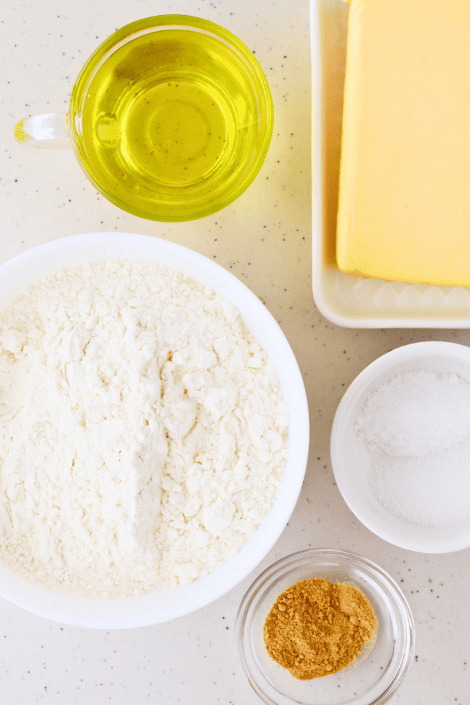 A top-down view of the key ingredients for making Puerto Rican arepas, including flour, butter, salt, garlic powder, and oil, showcasing the simple yet essential components of this traditional fried bread.