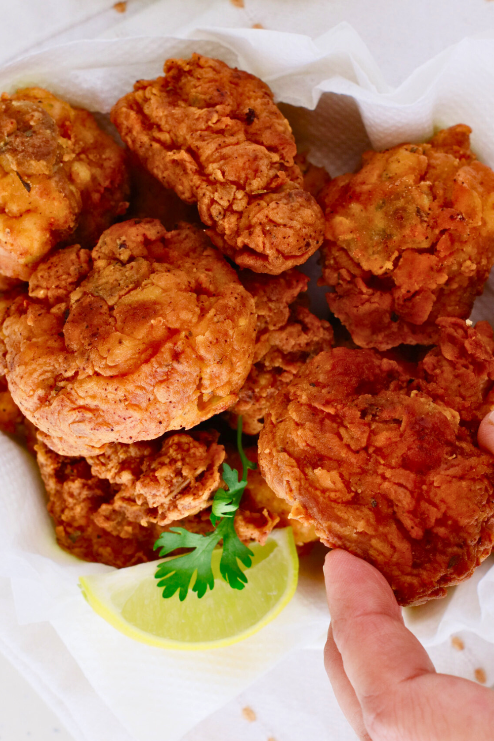 A basket of crispy chicharrones de pollo, Puerto Rican fried chicken bites, with a hand picking up a piece, garnished with fresh cilantro and lime wedges.