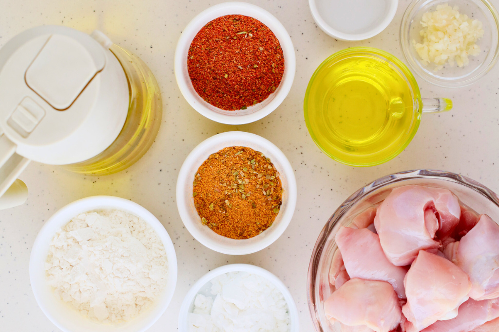 A top-down view of fresh ingredients for chicharrones de pollo, including chicken thighs, adobo and sazon seasoning, flour, cornstarch, garlic, and oil.